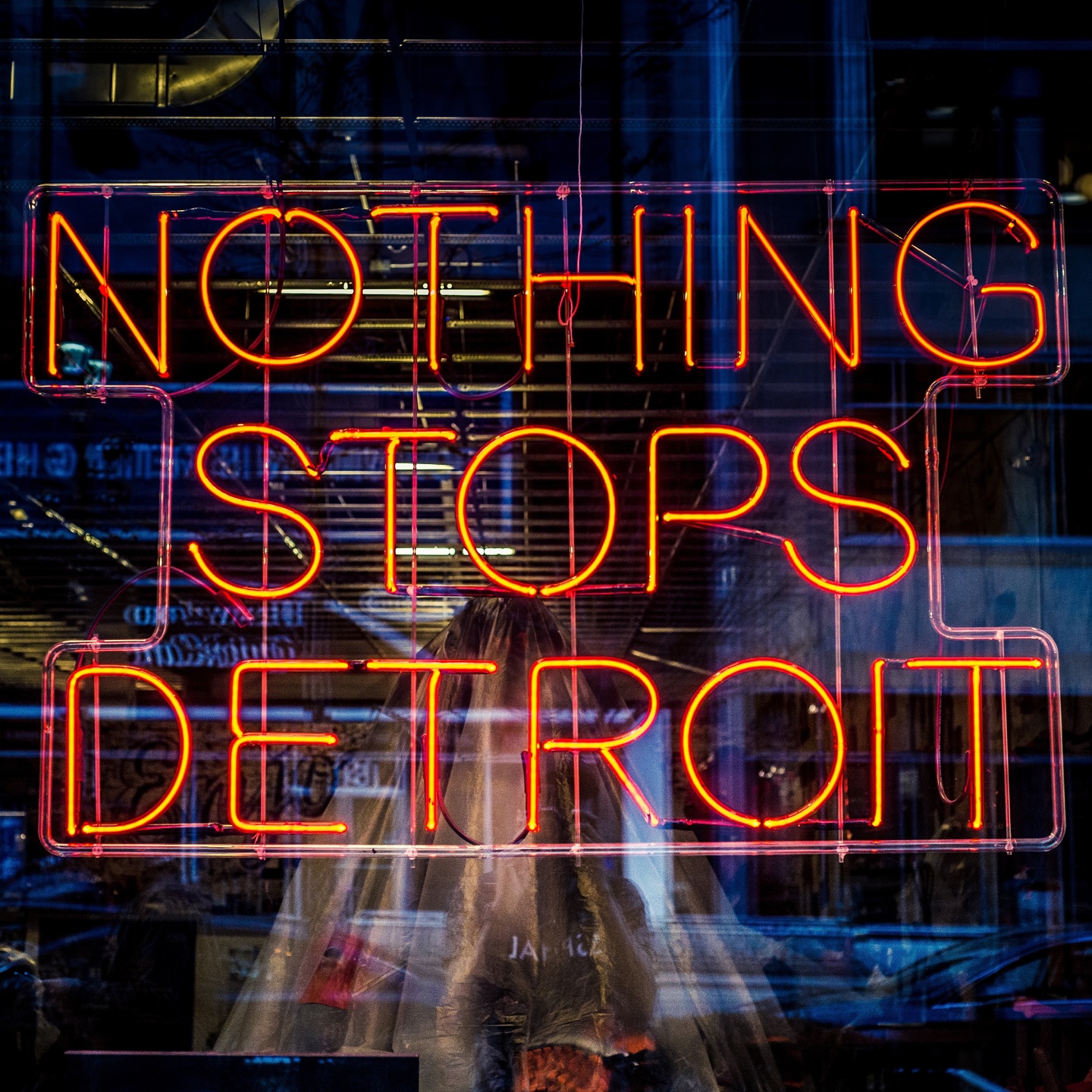 A photograph of an orange-colored neon light sign that reads “Nothing Stops Detroit” in all upper-case lettering. The neon sign is behind a translucent window.