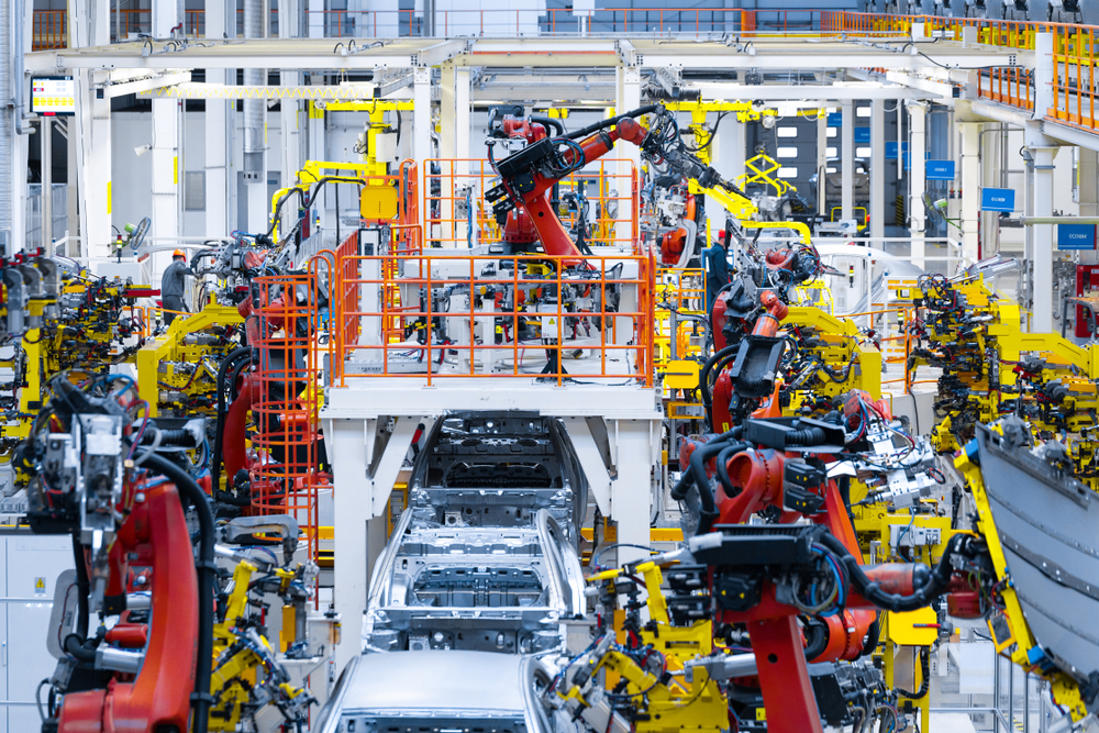 A photograph of an automotive body-in-white line in a modern automotive plant. The robots are yellow and red.